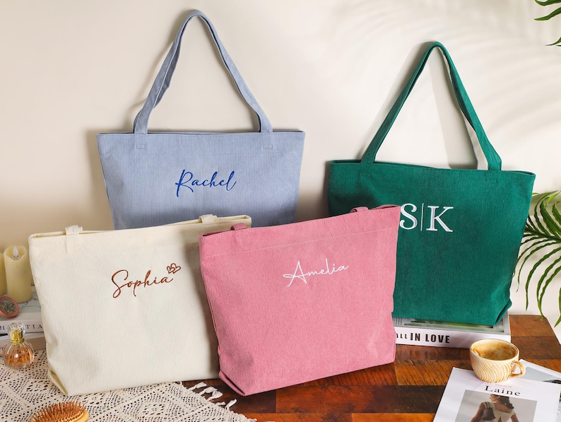 May include: Four personalized tote bags in various colors: light blue, green, pink, and off-white. Each bag features a name or initials embroidered in a contrasting color. The bags have long handles for carrying and are displayed on a wooden surface.