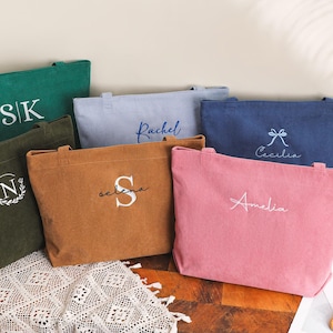 May include: A collection of corduroy tote bags in various colors, including green, brown, pink, and blue. Each bag features personalized embroidery with initials or names. The bags are rectangular with handles, suitable for carrying items.