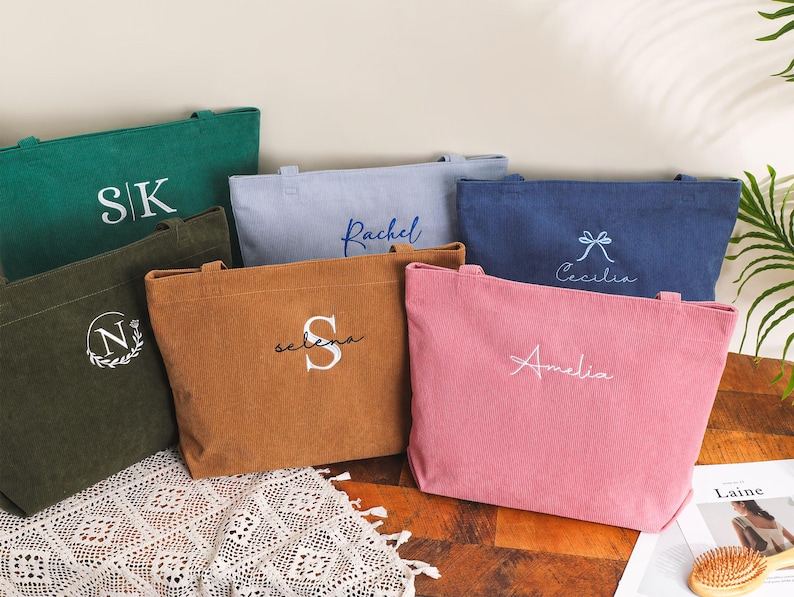 May include: A collection of corduroy tote bags in various colors, including green, brown, pink, and blue. Each bag features personalized embroidery with initials or names. The bags are rectangular with handles, suitable for carrying items.