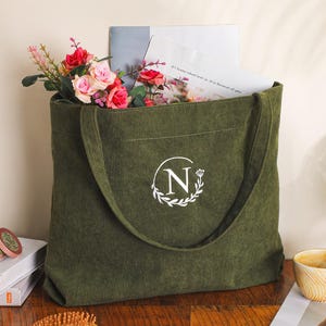 May include: Olive green corduroy tote bag with a white monogram "N" inside a laurel wreath. The bag is filled with flowers, papers, and a book. A small candle and a cup of coffee are next to the bag.