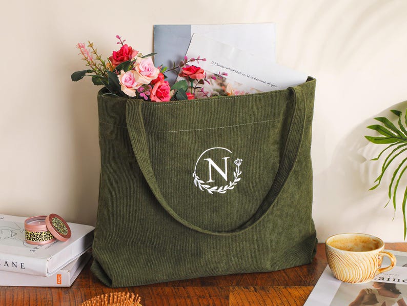 May include: Olive green corduroy tote bag with a white monogram "N" inside a laurel wreath. The bag is filled with flowers, papers, and a book. A small candle and a cup of coffee are next to the bag.
