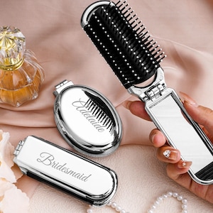 Peut inclure: Miroir compact argenté, peigne et flacon de parfum sur une surface rose. Le peigne est noir et argenté, et le miroir porte le nom "Adelaide". Un miroir compact avec le mot "Bridesmaid" est également visible.