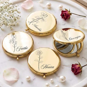 Pode incluir: Quatro espelhos de bolso redondos em metal dourado com tampas articuladas. Cada espelho apresenta uma ilustração floral de linhas pretas e um nome gravado em letra cursiva: Amelia, Faithe, Georgia e Monica. Um espelho está aberto.