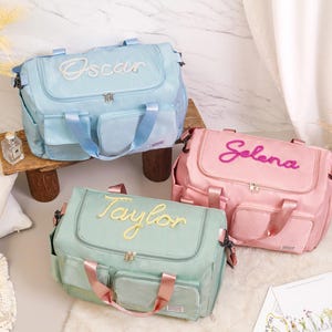 May include: Three personalized baby bags in pastel colors: light blue, pink, and mint green. Each bag has a name embroidered on the front: "Oscar", "Selena", and "Taylor". The bags have multiple pockets and handles, ideal for carrying baby essentials.