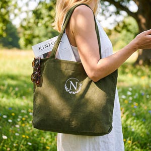 May include: Olive green corduroy tote bag with a white embroidered initial and laurel wreath design. The bag contains a magazine and sunglasses. The tote bag has long straps and is carried outdoors in a field.