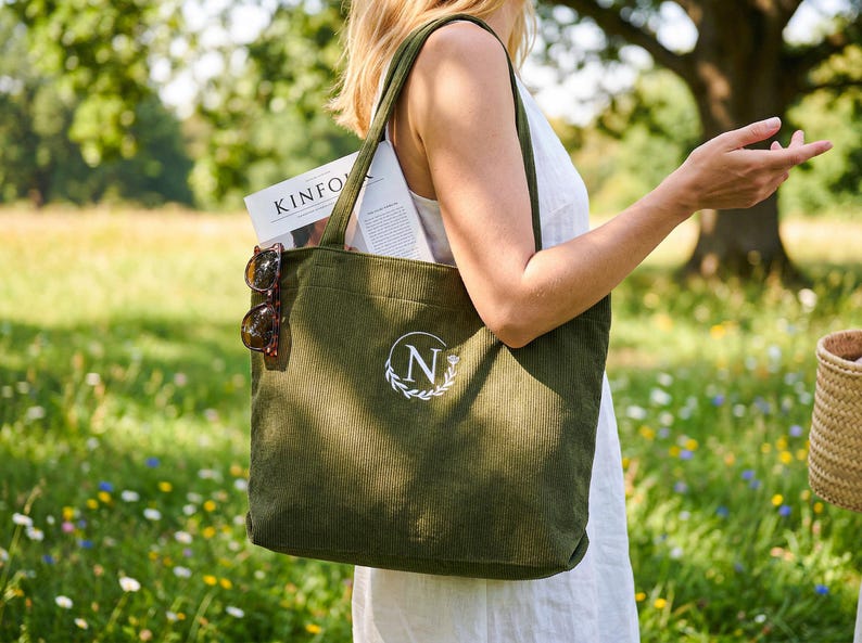 May include: Olive green corduroy tote bag with a white embroidered initial and laurel wreath design. The bag contains a magazine and sunglasses. The tote bag has long straps and is carried outdoors in a field.