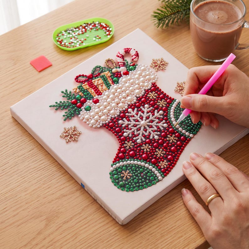 Puede incluir: Un kit de pintura de diamantes sobre lienzo cuadrado con un dise&ntilde;o de calcet&iacute;n navide&ntilde;o lleno de regalos, un bast&oacute;n de caramelo y copos de nieve, creado con cuentas rojas, verdes, blancas y doradas. Se usa un aplicador rosa para colocar las cuentas.