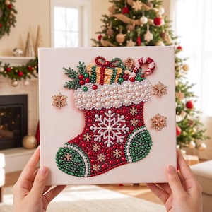 Puede incluir: Un lienzo cuadrado con un dise&ntilde;o de calcet&iacute;n navide&ntilde;o hecho con cuentas en rojo, verde y blanco. El calcet&iacute;n est&aacute; decorado con cuentas nacaradas, un copo de nieve blanco central, y un regalo, un bast&oacute;n de caramelo y ramas de pino.