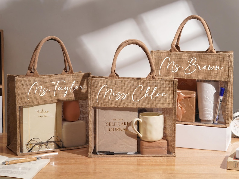 May include: Three rectangular jute tote bags with clear plastic windows on the front. Each bag features a white script name: Ms. Taylor, Miss Chloe, and Ms. Brown. The bags contain various items including journals, mugs, and small boxes.