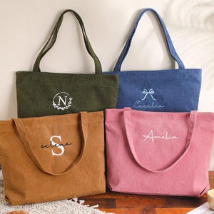 May include: Four corduroy tote bags in olive green, denim blue, tan, and pink. Each bag has a long handle and embroidered text: "N", "Cecilia", "Selena", and "Amelia". The bags are displayed on a wooden surface.