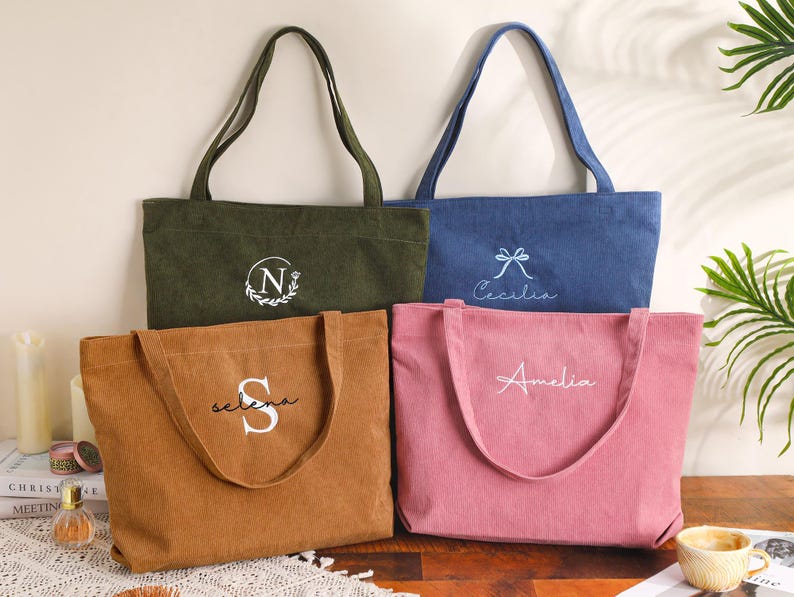 May include: Four corduroy tote bags in olive green, denim blue, tan, and pink. Each bag has a long handle and embroidered text: "N", "Cecilia", "Selena", and "Amelia". The bags are displayed on a wooden surface.