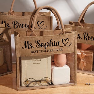 May include: A set of rectangular jute tote bags with two handles and a clear plastic window on the front. Each bag features black text, including names like Ms. Sophia, Ms. Brown, and Ms. Reeves, along with the phrase BEST TEACHER EVER and a heart icon.