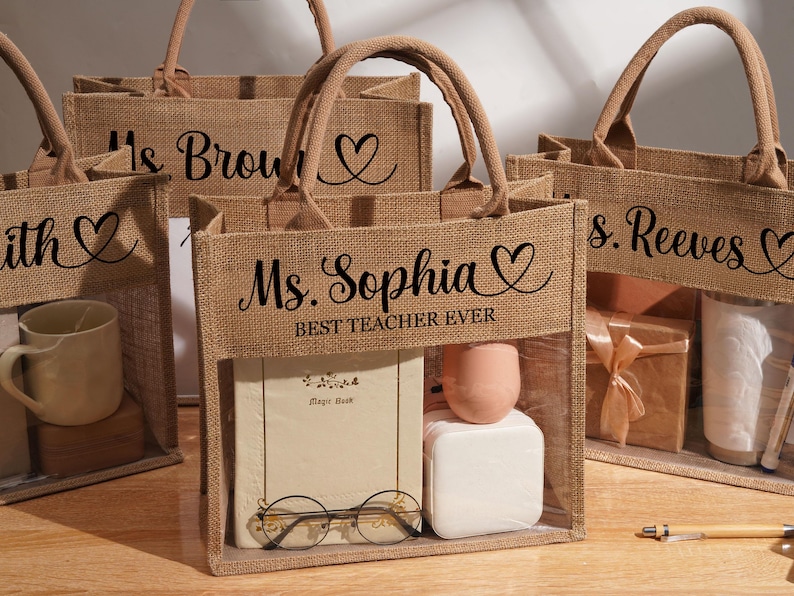 May include: A set of rectangular jute tote bags with two handles and a clear plastic window on the front. Each bag features black text, including names like Ms. Sophia, Ms. Brown, and Ms. Reeves, along with the phrase BEST TEACHER EVER and a heart icon.