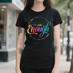 May include: Black crew neck t-shirt with a colorful graphic that reads "YOU ARE Enough". The design includes words like "Kind", "Tough", "Powerful", "Loved", "Valued", "Strong", "Bold", "Brave", "Smart", and "Capable".