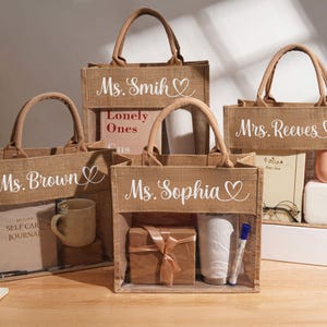 May include: Four rectangular jute tote bags with brown fabric handles and a clear plastic window on the front. Each bag features white cursive text and a heart icon, personalized with the names Ms. Brown, Ms. Smith, Mrs. Reeves, and Ms. Sophia.