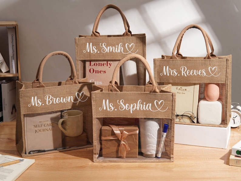 May include: Four rectangular jute tote bags with brown fabric handles and a clear plastic window on the front. Each bag features white cursive text and a heart icon, personalized with the names Ms. Brown, Ms. Smith, Mrs. Reeves, and Ms. Sophia.