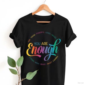 May include: Black t-shirt with the words "You Are Enough" in a colorful, rainbow-style script. The design includes words like "Kind," "Strong," and "Brave" in a circular pattern around the main text. The shirt is hanging on a wooden hanger.