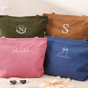 May include: Four corduroy tote bags in olive green, brown, pink, and blue. Each bag has a personalized embroidered design: a floral initial, a name, and a bow with a name. The bags are on a cream-colored surface.