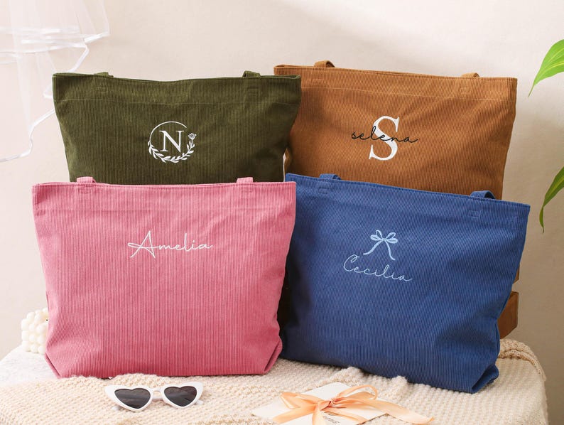 May include: Four corduroy tote bags in olive green, brown, pink, and blue. Each bag has a personalized embroidered design: a floral initial, a name, and a bow with a name. The bags are on a cream-colored surface.
