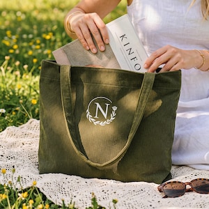May include: Olive green corduroy tote bag with a white monogram and the letter "N" inside a wreath. The bag has long straps and is sitting on a white lace blanket. A book with the word "KINFO" is being placed inside.