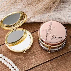 May include: A gold-toned, open compact mirror next to a stack of rose gold, silver, and gold compact mirrors. The top mirror has a floral design and the name "Georgia". Makeup brushes and a pearl necklace are also present.
