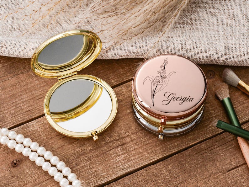 May include: A gold-toned, open compact mirror next to a stack of rose gold, silver, and gold compact mirrors. The top mirror has a floral design and the name "Georgia". Makeup brushes and a pearl necklace are also present.