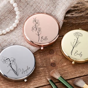 May include: Three personalized compact mirrors in rose gold, silver, and gold. Each mirror features a floral design and a name: Belle, Angela, and Emily. Makeup brushes and a pearl necklace are also visible.