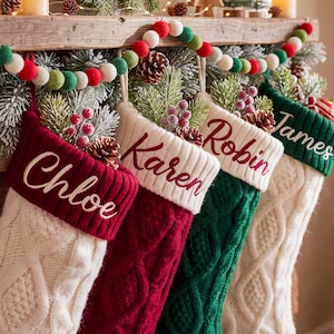 May include: Four personalized Christmas stockings with cable knit patterns, featuring the names Chloe, Karen, Robin, and James embroidered in red or white. The stockings are decorated with pine sprigs, pinecones, and red berries.