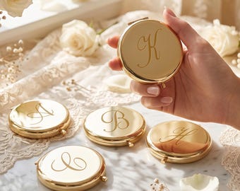 Puede incluir: Una colección de espejos de bolsillo redondos de metal dorado, cada uno con una letra cursiva grabada en la tapa. Las letras A, B, C, H y K son visibles en los espejos individuales, que tienen una superficie pulida y reflectante.
