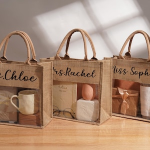 May include: Three rectangular jute tote bags with tan fabric handles and a clear plastic front window. Each bag features black cursive text: Ms. Chloe, Mrs. Rachel, and Miss Sophia. The bags contain various items including a mug, notebook, and gifts.
