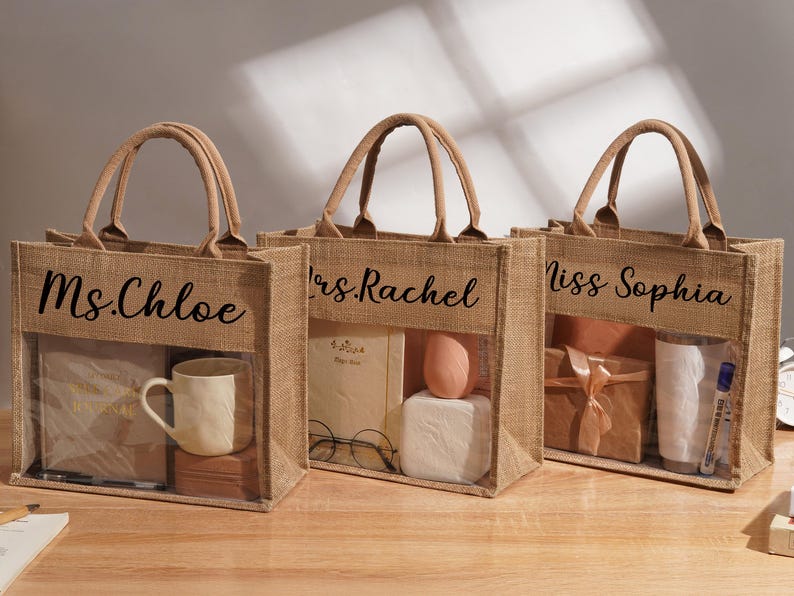 May include: Three rectangular jute tote bags with tan fabric handles and a clear plastic front window. Each bag features black cursive text: Ms. Chloe, Mrs. Rachel, and Miss Sophia. The bags contain various items including a mug, notebook, and gifts.