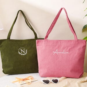 May include: Two corduroy tote bags. One is olive green with a white embroidered "N" inside a wreath. The other is pink with the name "Amelia" embroidered in white. Both have long handles and are sitting on a cream-colored surface.
