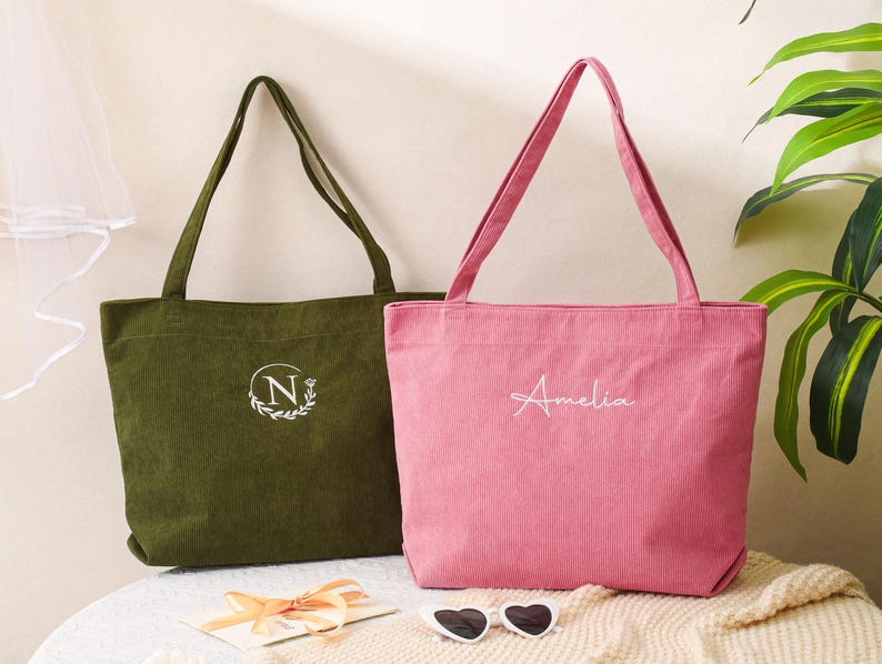 May include: Two corduroy tote bags. One is olive green with a white embroidered "N" inside a wreath. The other is pink with the name "Amelia" embroidered in white. Both have long handles and are sitting on a cream-colored surface.
