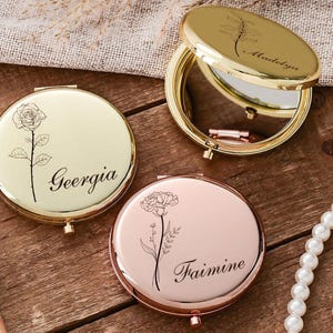 May include: Three personalized compact mirrors in gold and rose gold tones are displayed on a wooden surface. Each mirror features a floral design and a name: "Geergia", "Madelyn", and "Faimine". A makeup brush and a pearl necklace are also visible.