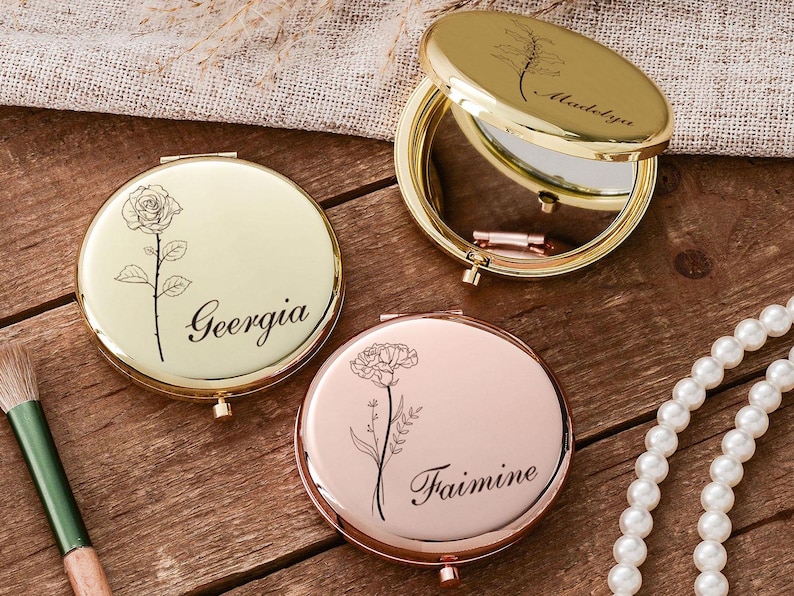 May include: Three personalized compact mirrors in gold and rose gold tones are displayed on a wooden surface. Each mirror features a floral design and a name: "Geergia", "Madelyn", and "Faimine". A makeup brush and a pearl necklace are also visible.