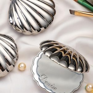 Pode incluir: Três espelhos de bolso de metal prateado em forma de concha. Um espelho está aberto, revelando uma superfície refletora com o nome Denise gravado em letra cursiva. O exterior de cada espelho apresenta um design de concha canelada.