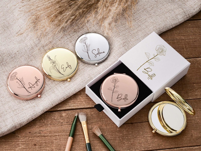 May include: A collection of personalized compact mirrors in various colors, including rose gold, silver, and gold. Each mirror features a floral design and a name. One mirror is in a white box with a rose illustration.