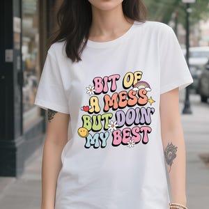 Może przedstawiać: Biały t-shirt z napisem "Bit of a Mess But Doin' My Best" w kolorowej, retro czcionce. Projekt zawiera tęczę, gwiazdy, kwiaty i uśmiechniętą buzię. Codzienny, swobodny t-shirt.