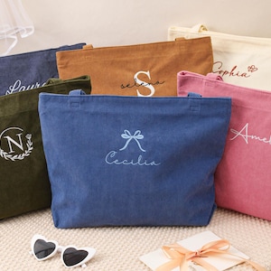 May include: A collection of corduroy tote bags in various colors, including navy, olive green, brown, cream, pink, and blue. Each bag features personalized embroidery with names or initials. Heart-shaped sunglasses and a gift card are in the foreground.