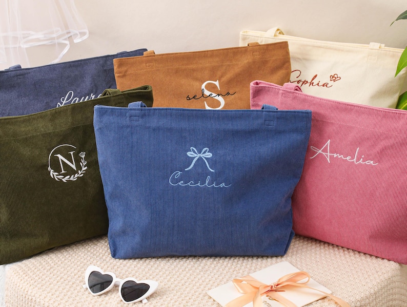 May include: A collection of corduroy tote bags in various colors, including navy, olive green, brown, cream, pink, and blue. Each bag features personalized embroidery with names or initials. Heart-shaped sunglasses and a gift card are in the foreground.