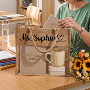 May include: A rectangular burlap tote bag with two fabric handles, featuring the black printed text Ms. Sophia and a heart icon. The bag has a clear front panel displaying a beige journal titled My Daily Self Care Journal, a cream mug, and a small box.