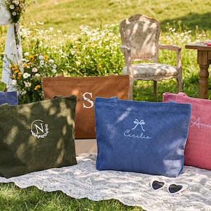 May include: An outdoor scene features several canvas tote bags in various colors: blue, green, brown, and pink. Each bag has a personalized design, including names and initials. The bags are arranged on a white lace cloth, with a vintage chair and table in the background.