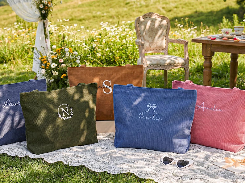 May include: An outdoor scene features several canvas tote bags in various colors: blue, green, brown, and pink. Each bag has a personalized design, including names and initials. The bags are arranged on a white lace cloth, with a vintage chair and table in the background.