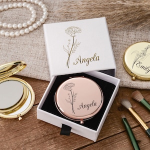 May include: A collection of personalized compact mirrors and a white gift box with floral designs and the name "Angela" in gold script. Makeup brushes and a pearl necklace are also visible.