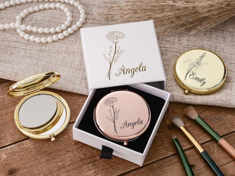 May include: A collection of personalized compact mirrors and a white gift box with floral designs and the name "Angela" in gold script. Makeup brushes and a pearl necklace are also visible.