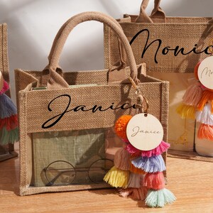 May include: Three tan burlap tote bags with clear plastic windows, each featuring a black name printed on the front. Each bag includes a round wooden tag with a name and a multi-colored tassel keychain in shades of pink, orange, yellow, and blue.