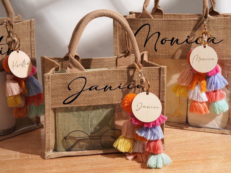 May include: Three tan burlap tote bags with clear plastic windows, each featuring a black name printed on the front. Each bag includes a round wooden tag with a name and a multi-colored tassel keychain in shades of pink, orange, yellow, and blue.