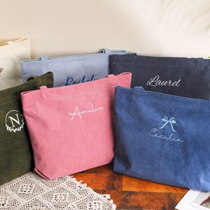 May include: A collection of corduroy tote bags in various colors, including pink, blue, and green. Each bag is personalized with embroidered names or initials in white or light blue thread. The bags are displayed on a table with a decorative lace runner.