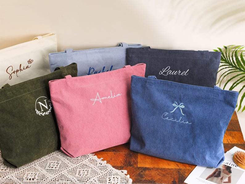May include: A collection of corduroy tote bags in various colors, including pink, blue, and green. Each bag is personalized with embroidered names or initials in white or light blue thread. The bags are displayed on a table with a decorative lace runner.