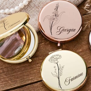 May include: A collection of personalized compact mirrors in gold, rose gold, and silver. Each mirror features a floral design and a name, such as "Arianna" and "Georgia", engraved on the surface. The open gold mirror reveals a reflective surface.